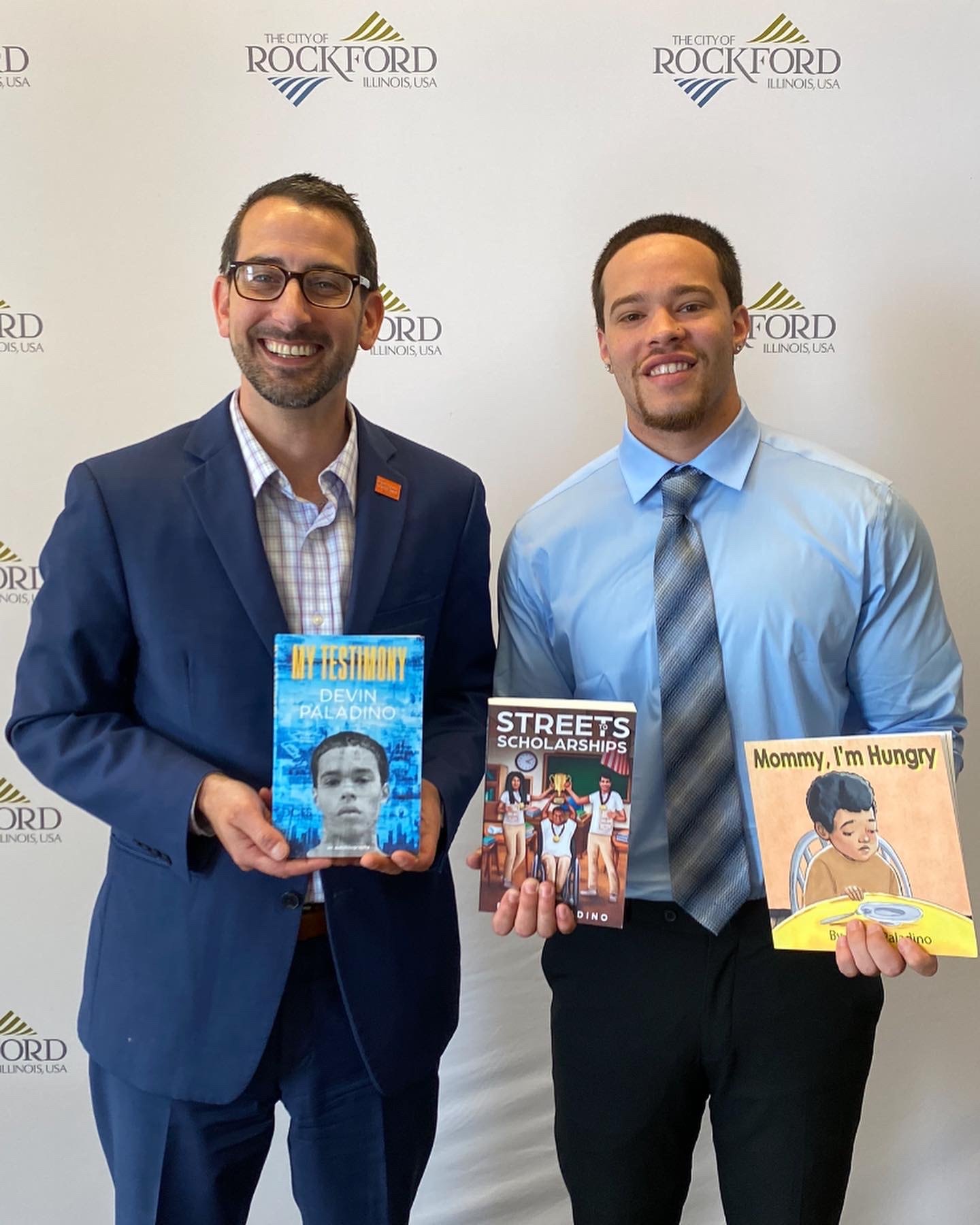 Devin Paladino holding three of his books beside a city official in front of a City of Rockford, Illinois backdrop