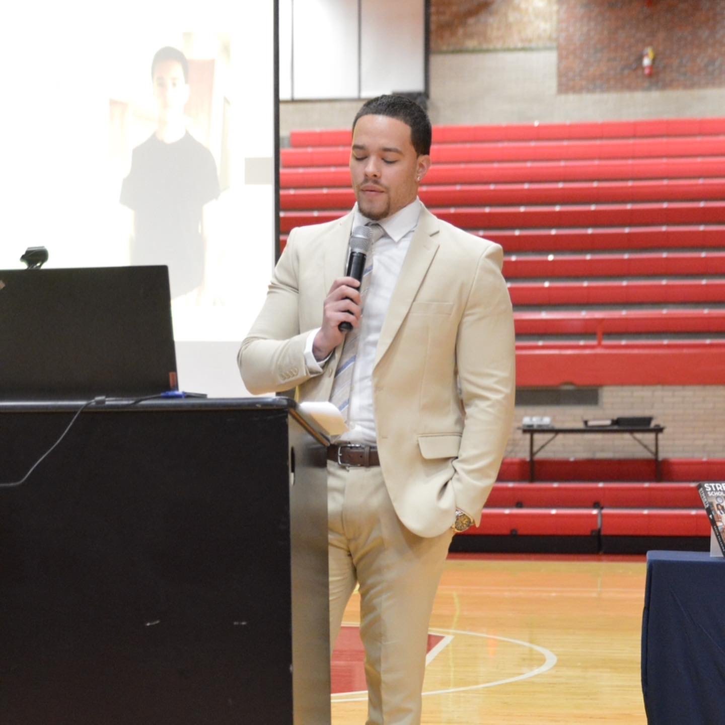 Devin Paladino speaking at a lectern in a school gymnasium