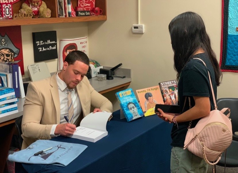 Devin Paladino in a suit signing a book for a reader at a bookstore table stacked with his titles