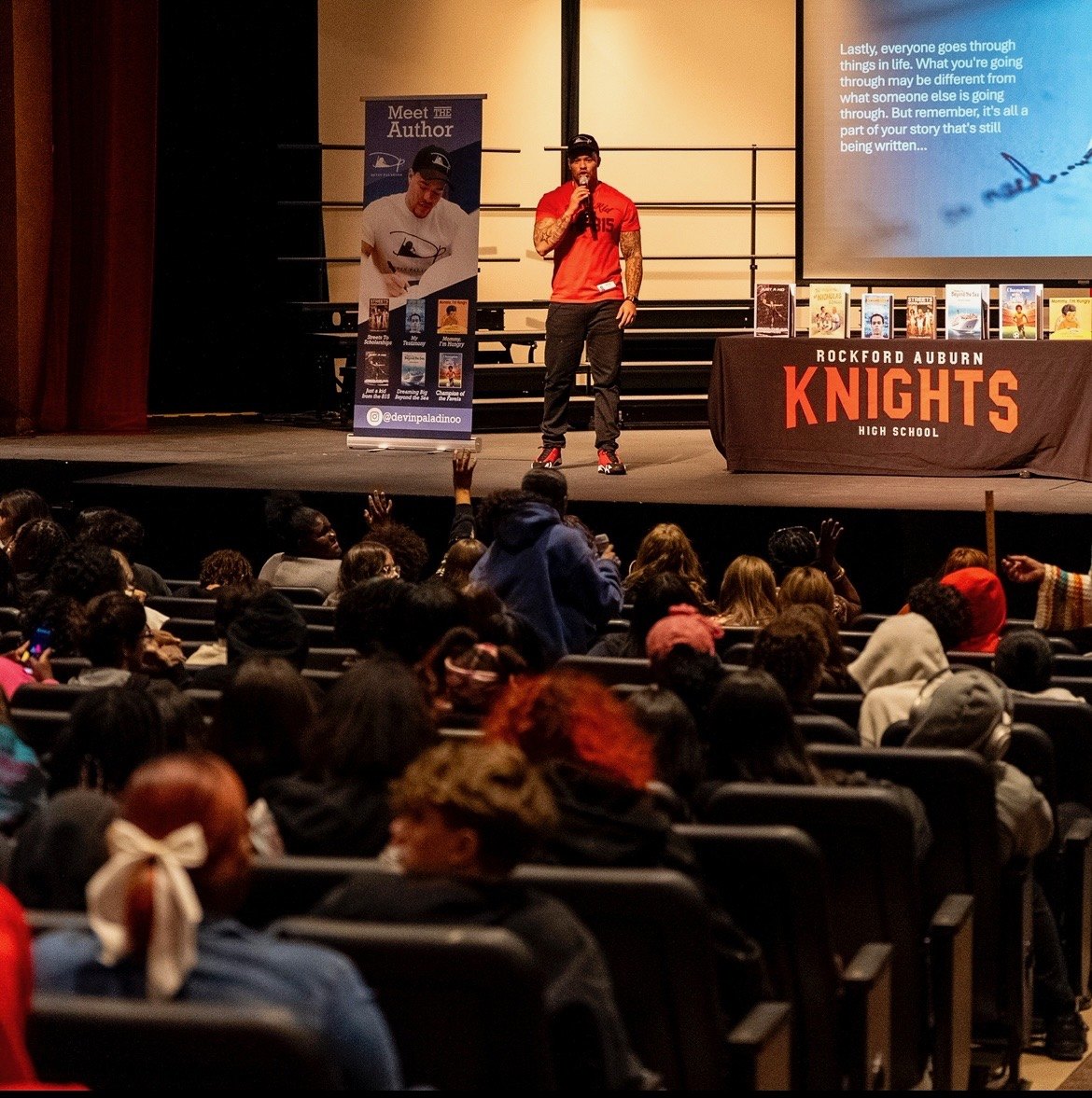 Devin Paladino on stage in a full high-school auditorium, a “Meet the Author” banner and a table of his books beside him